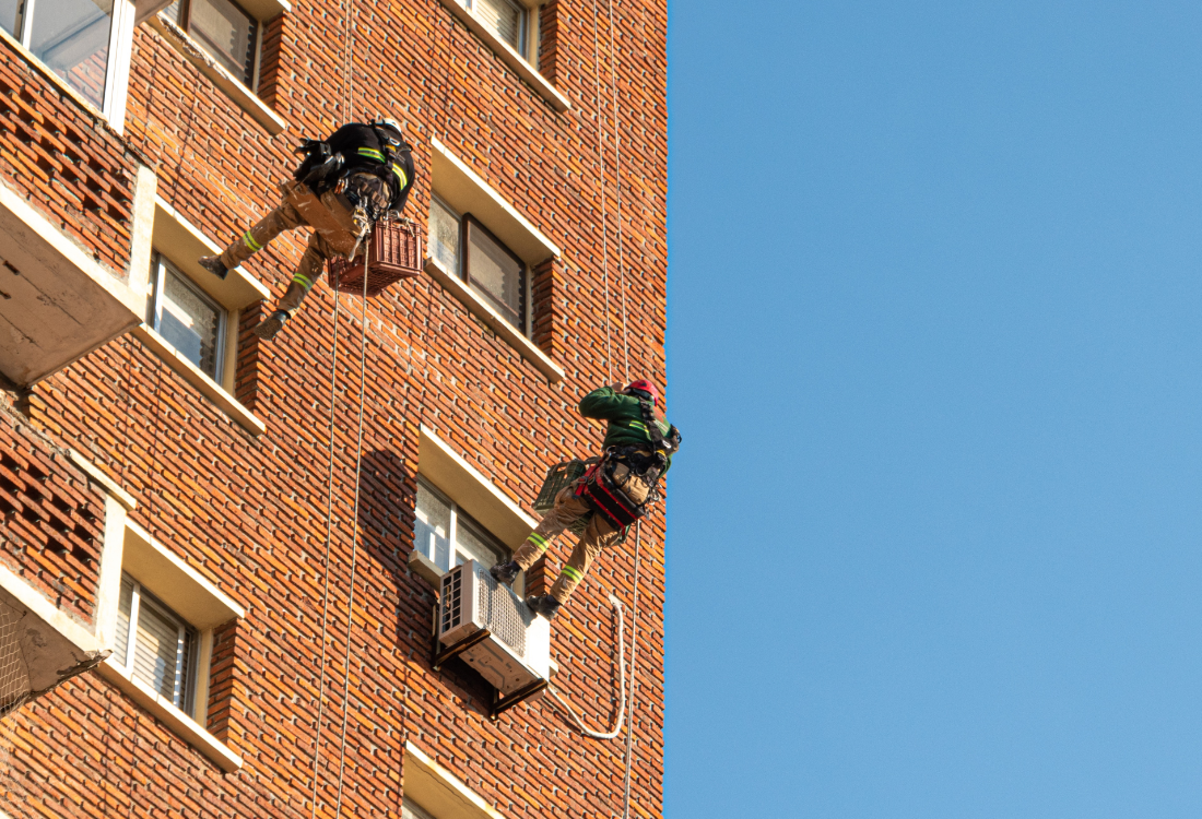 Workers using ropes and safety harnesses to perform maintenance at height on a tall brick building, demonstrating the need for safe bespoke access solutions.