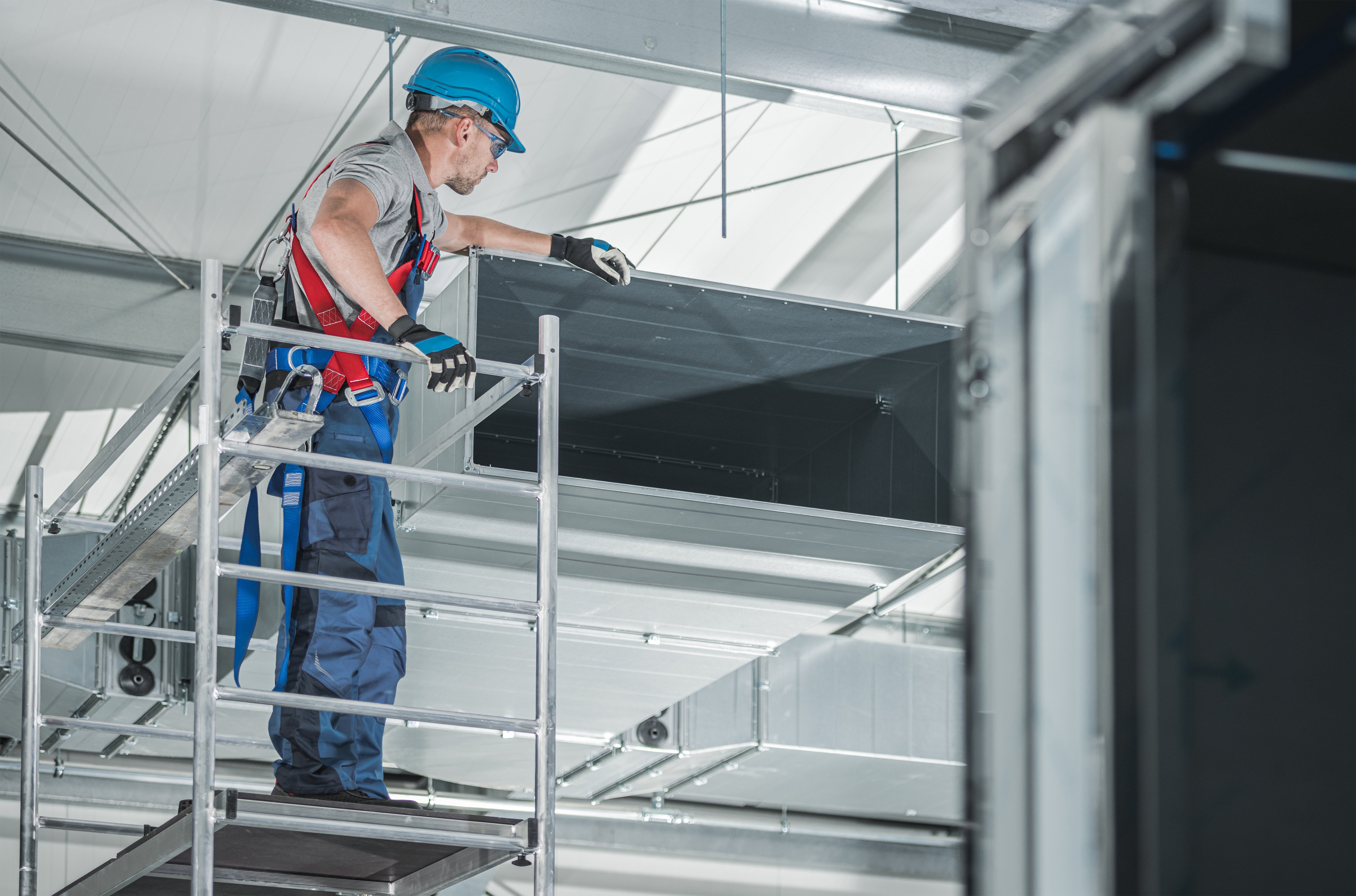 Worker using access equipment while inspecting overhead ductwork in a confined industrial space.