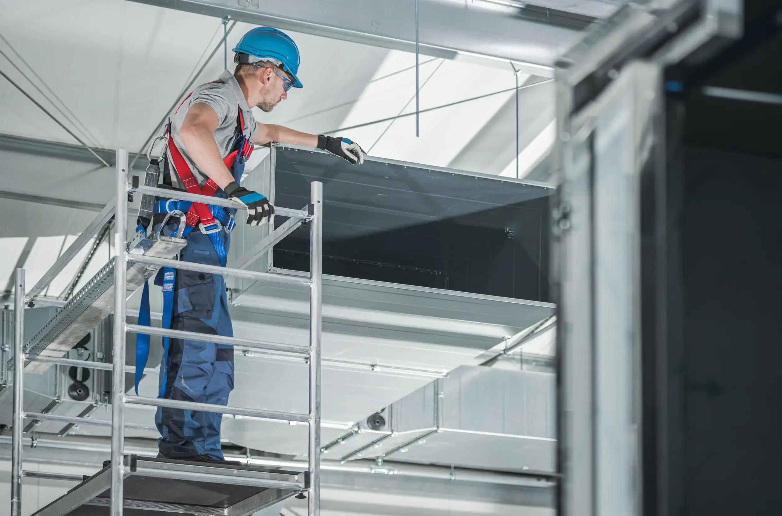 Worker using access equipment while inspecting overhead ductwork in a confined industrial space.